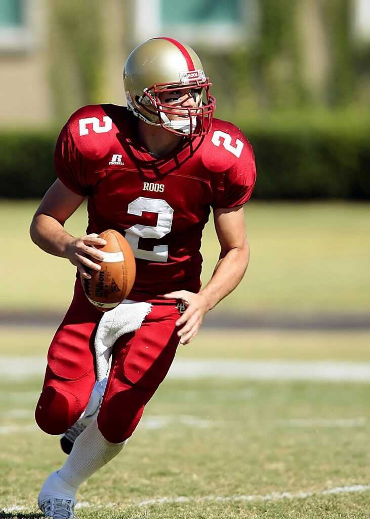 Quarterback in red jersey running with football on grassy field during daytime sporting event.